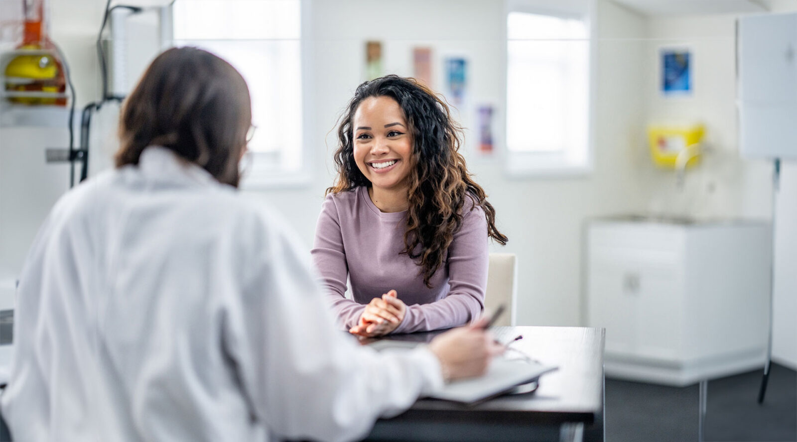 A young woman of mixed race, sits with her female doctor as they discuss her health concerns. The patient is dressed casually and has a smile on her face.